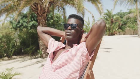 african american man sitting on deckchair wearing sunglasses, relaxing on sunny beach, slow motion