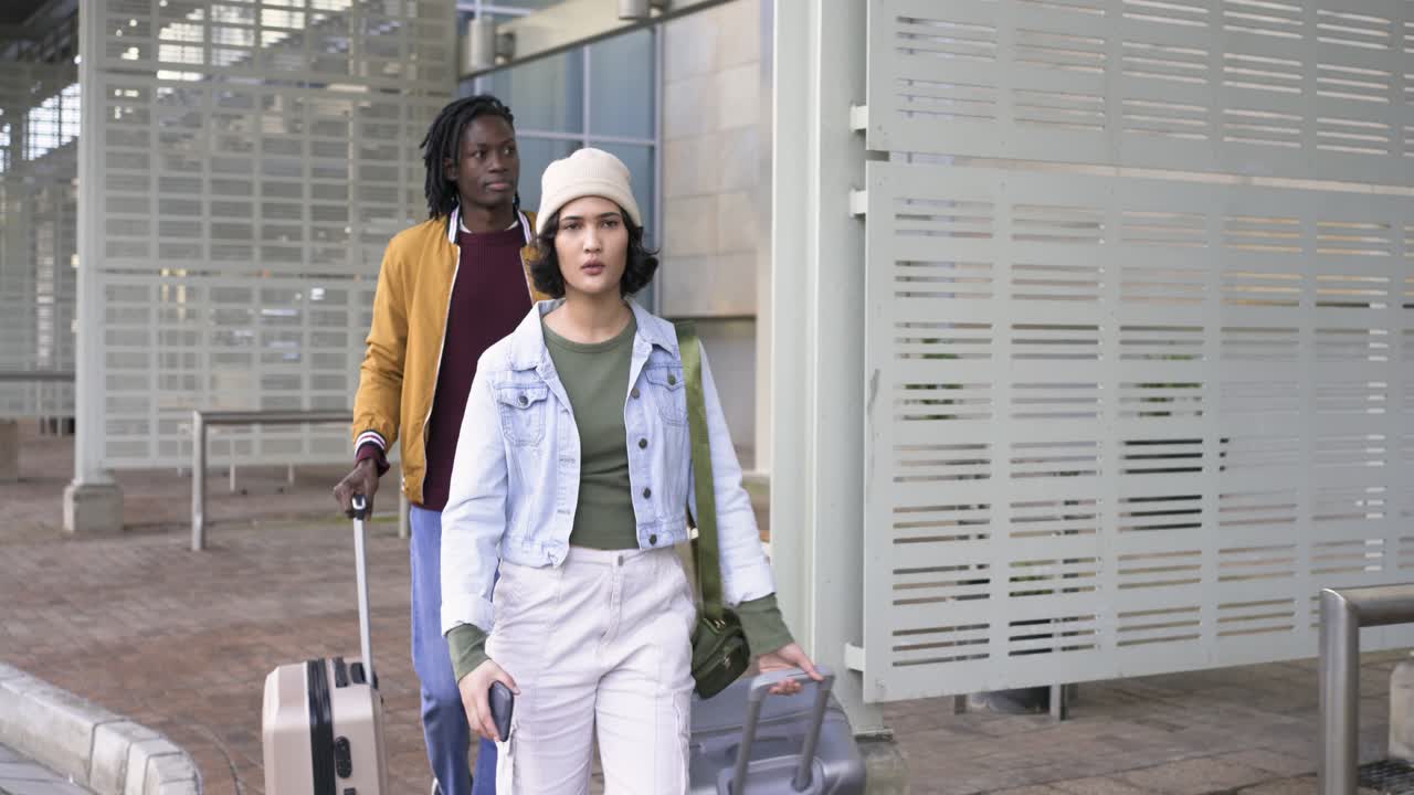 African American man and Asian woman travelers checking smartphone, rolling suitcases in courtyard