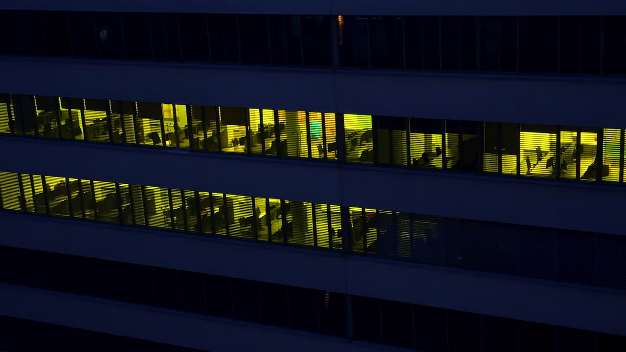 Multi-storied office building with desks and tables. Dark time footage outside the building with light switched on inside