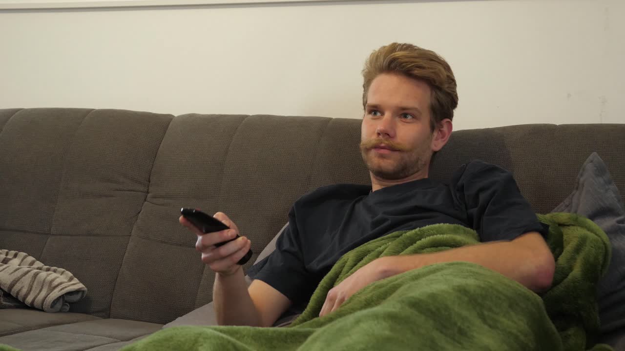 En blond fyr med et imponerende overskæg ligger på sofaen og zapper kanaler på tv'et.