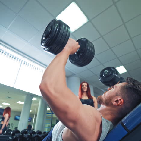side view of a strong man with biceps lying on desk and lifting dumbbells in gym. muscular man doing workout on the background of sport center. pretty woman looks at bodybuilders training.