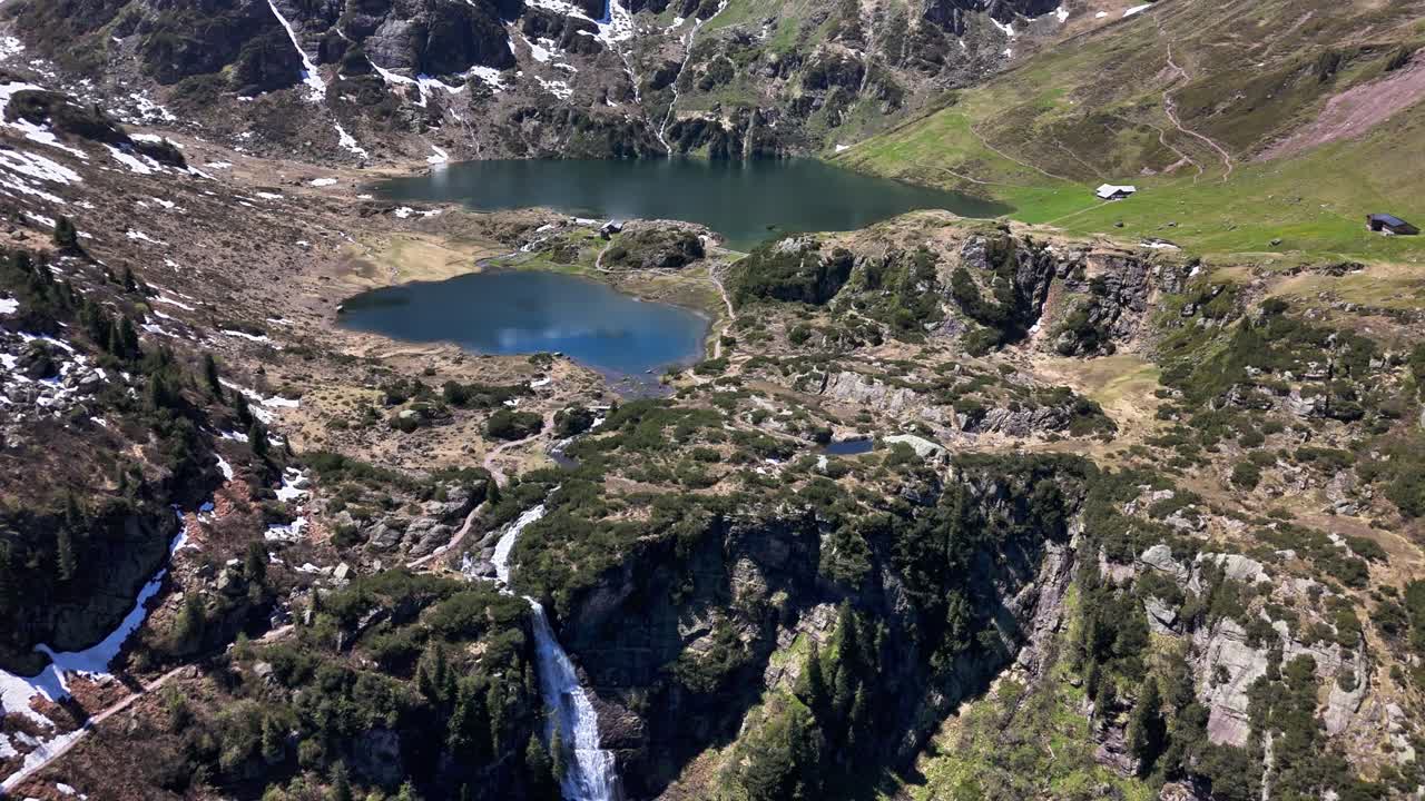 Tranquil Murgsee mirrors a vibrant alpine sky, surrounded by snow patches, lush green slopes and rocky outcrops and cascades, nestled in rugged Swiss mountains, drone pulling out