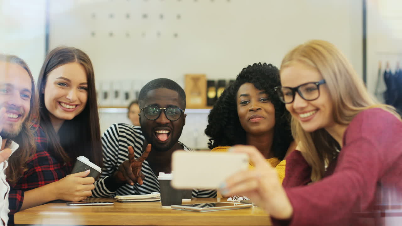Camera focuses on a multiethnic group of friends through the window making a selfie and doing funny gestures while they are sitting at a table in a cafe