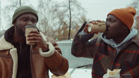 two black men having snack with coffee and chatting by car