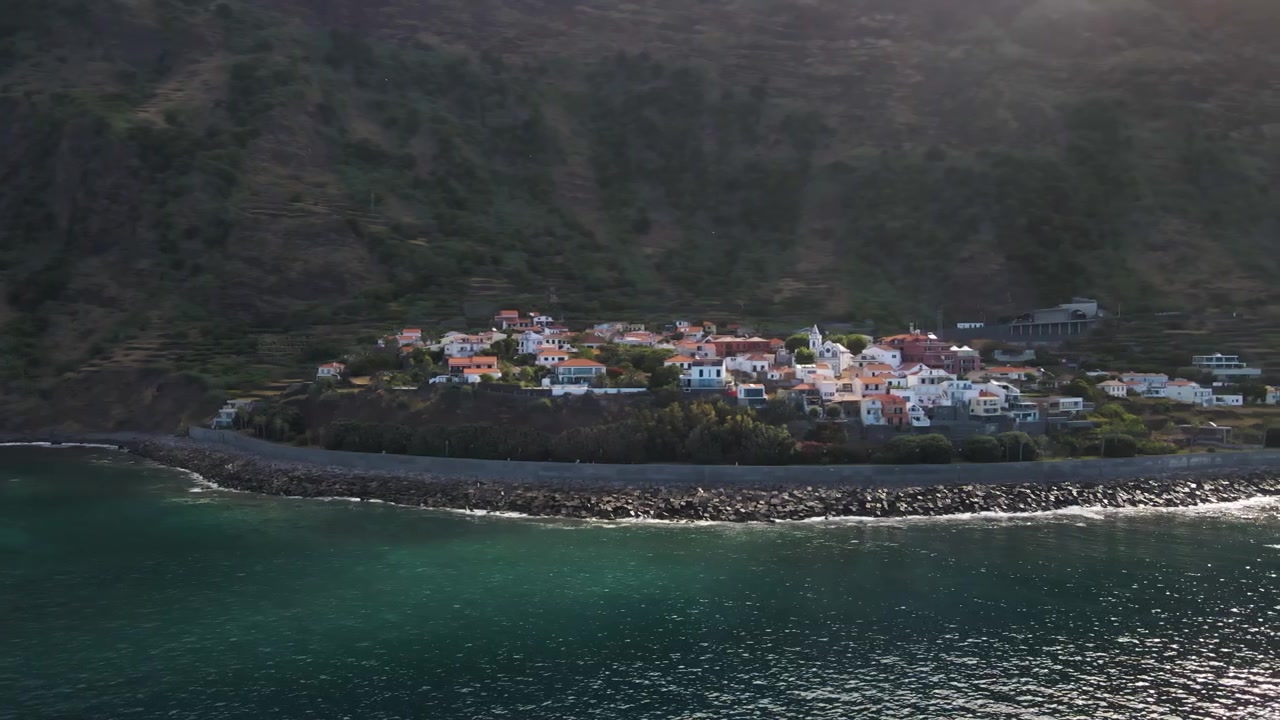 Riprese aeree che mostrano il pittoresco villaggio di Jardim do Mar, circondato da imponenti scogliere e lussureggianti terrazzamenti verdi. Il drone si muove verso destra, svelando il vasto orizzonte atlantico.