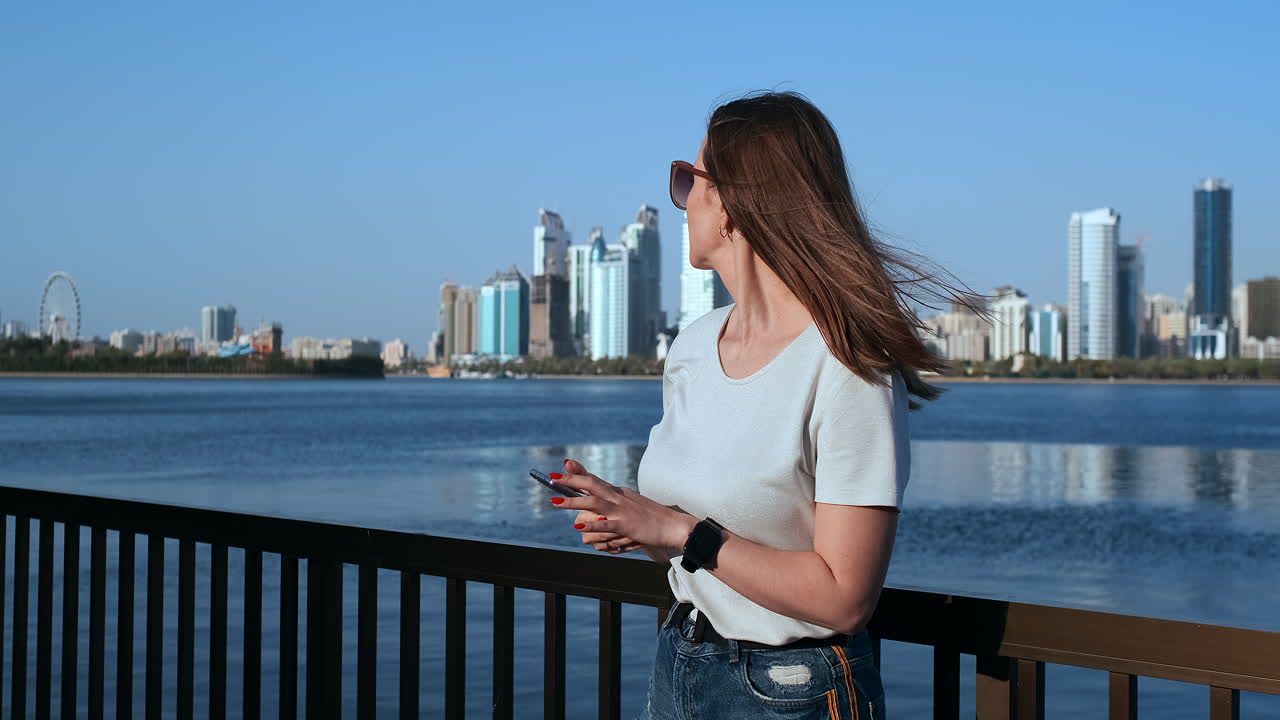 hermosa chica con cabello largo con gafas de sol usando una aplicación de teléfono inteligente en el muelle de sunset river cerca.