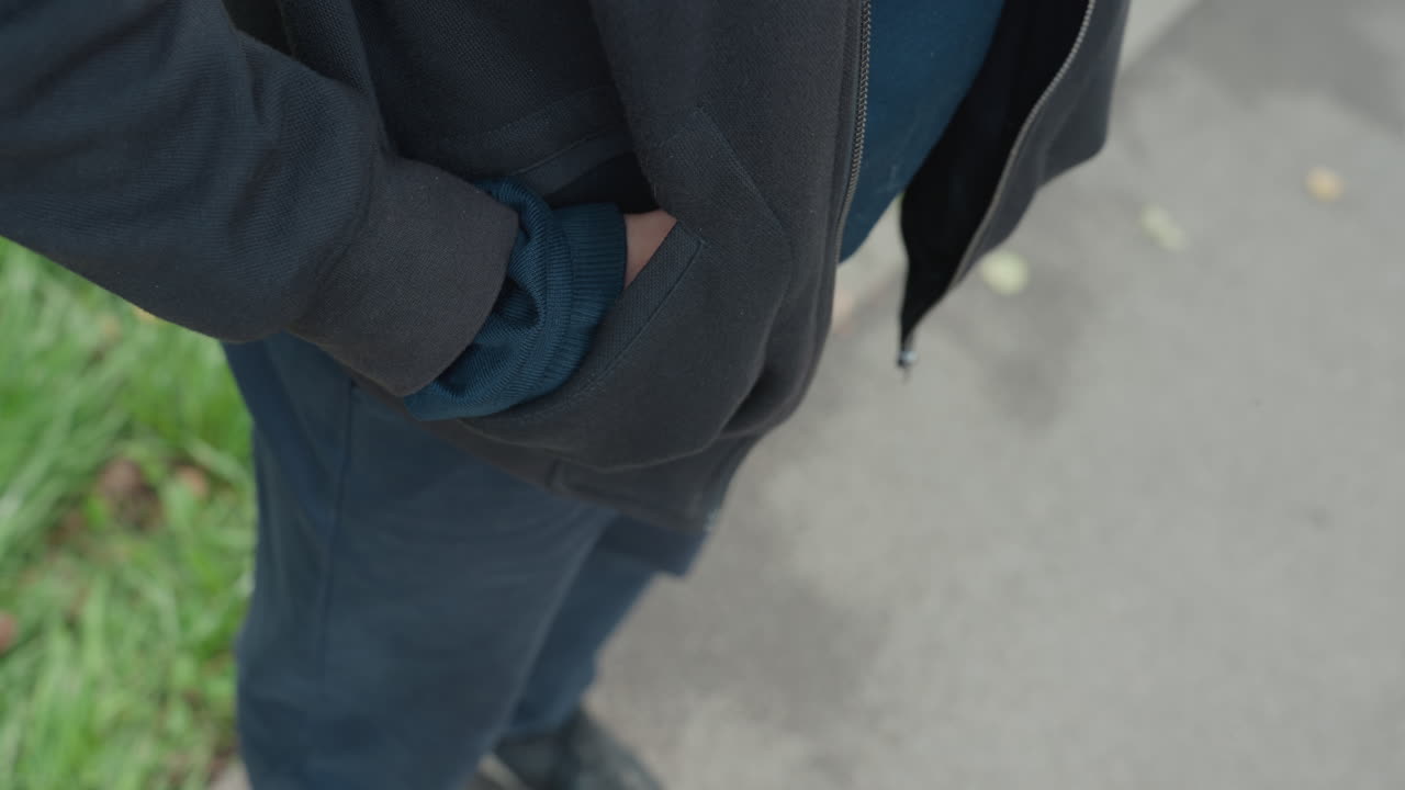 man fidgeting on pavement near grass, hands clasping and adjusting jacket, close topdown framing, sneakers and jeans visible, nervous anticipation, subtle motion and breath, urban walkway atmosphere
