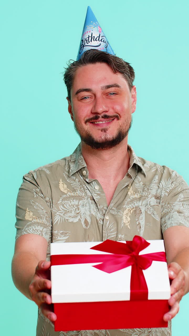 Uomo adulto con cappellino da festa che sorride felice mentre offre un regalo di compleanno a qualcuno che festeggia un evento, generosità