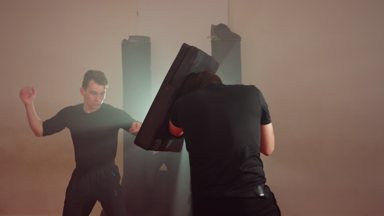 Karate practitioner training in gym during sparring exercise, man in fighting stance preparing to strike while partner holds shield for defense, intense practice on red mats with punching bags in background