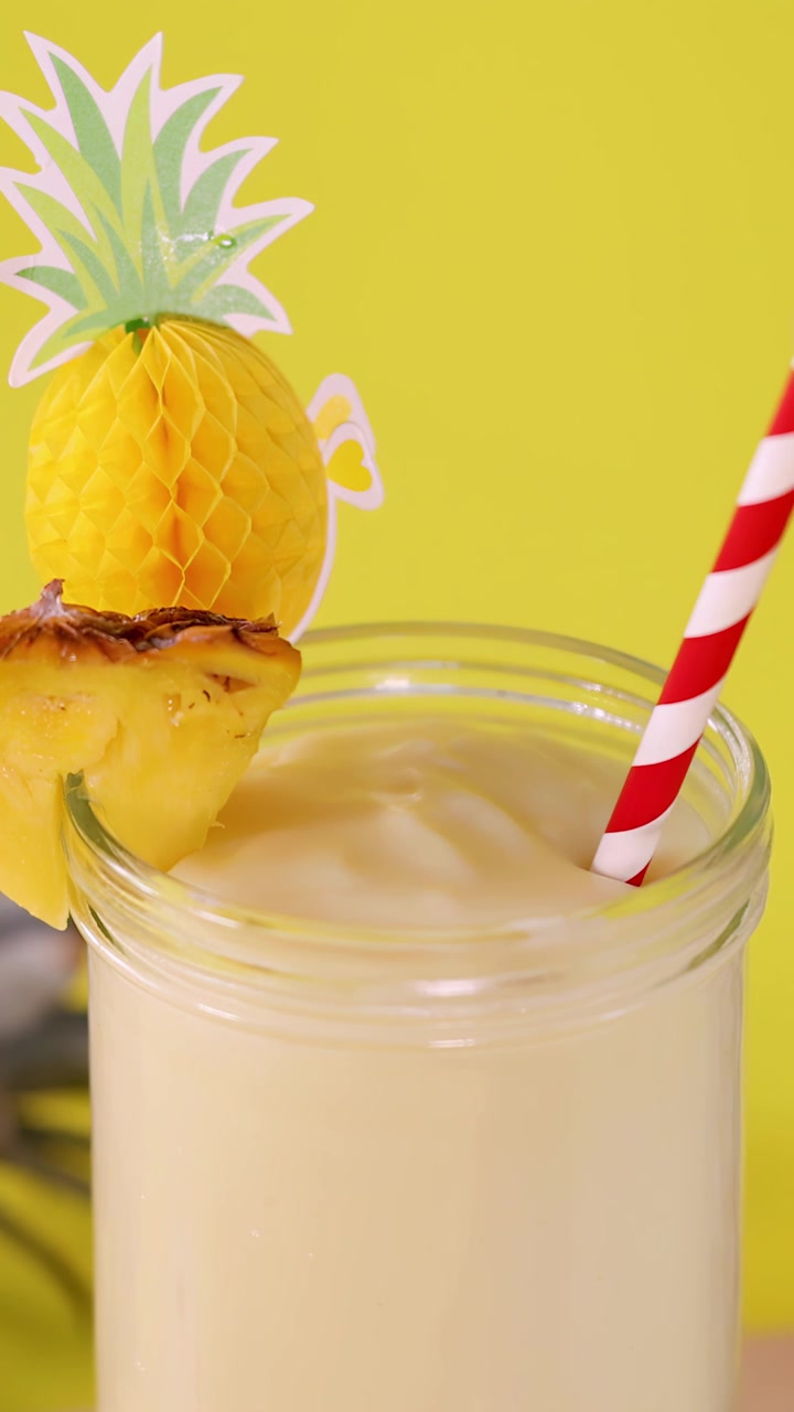 Primo piano di un frullato cremoso all'ananas in un bicchiere, una mano mescola una cannuccia a strisce su uno sfondo giallo brillante