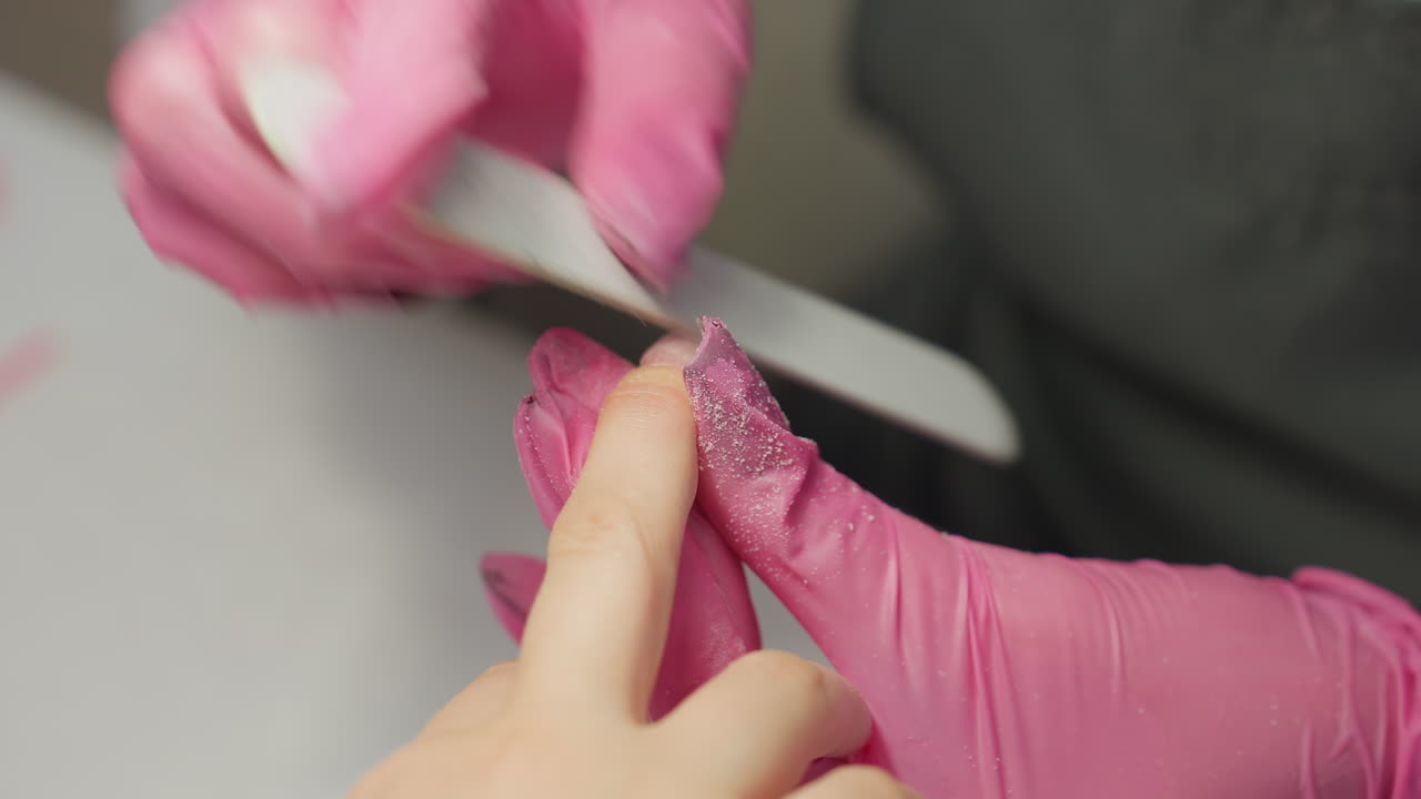 Nail technician in pink gloves filing client's fingernail while fine particles from nail shaping accumulate visibly on gloves, emphasizing detailed work and cleanliness in nail salon environment