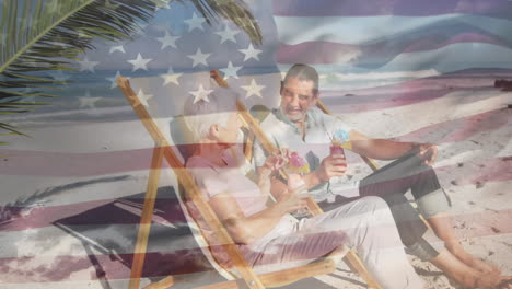 american flag waving against senior couple enjoying drinks sitting on deck chair at the beach
