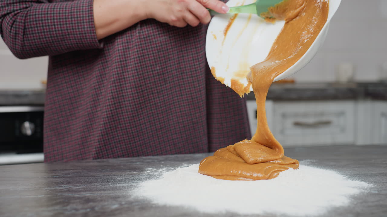 Partial view of woman pouring thick caramel-colored batter into flour on countertop using green spatula, smooth, creamy mixture flows from white bowl