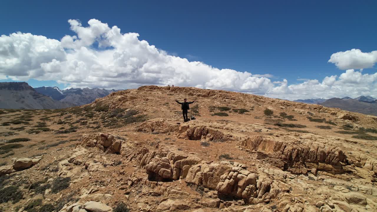 joven parado en el borde del acantilado y levantando las manos en la cima de altas montañas rocosas en un día soleado en el valle spiti india, aéreo