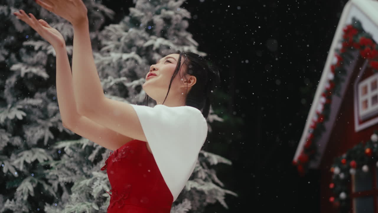 A woman in a red dress and white shawl catches snow outdoors by a festive house