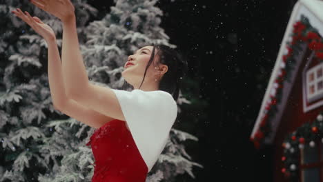 a woman in a red dress and white shawl catches snow outdoors by a festive house