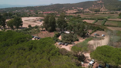 Vista-Aérea-Del-Pinar-Mediterráneo-Con-Estacionamiento-De-Autos-Cerca-De-La-Playa-Con-Montañas-En-El-Fondo-De-Cerdeña,-Italia---Retroceso-De-Tiro-De-Drones