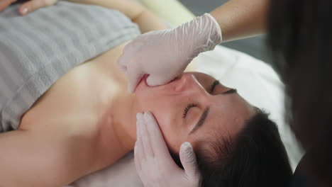 High angle of individual lying on massage chair closes eyes gently as beauty expert wearing safety gloves dips fingernail inside mouth while other hand massages cheeks with professional care