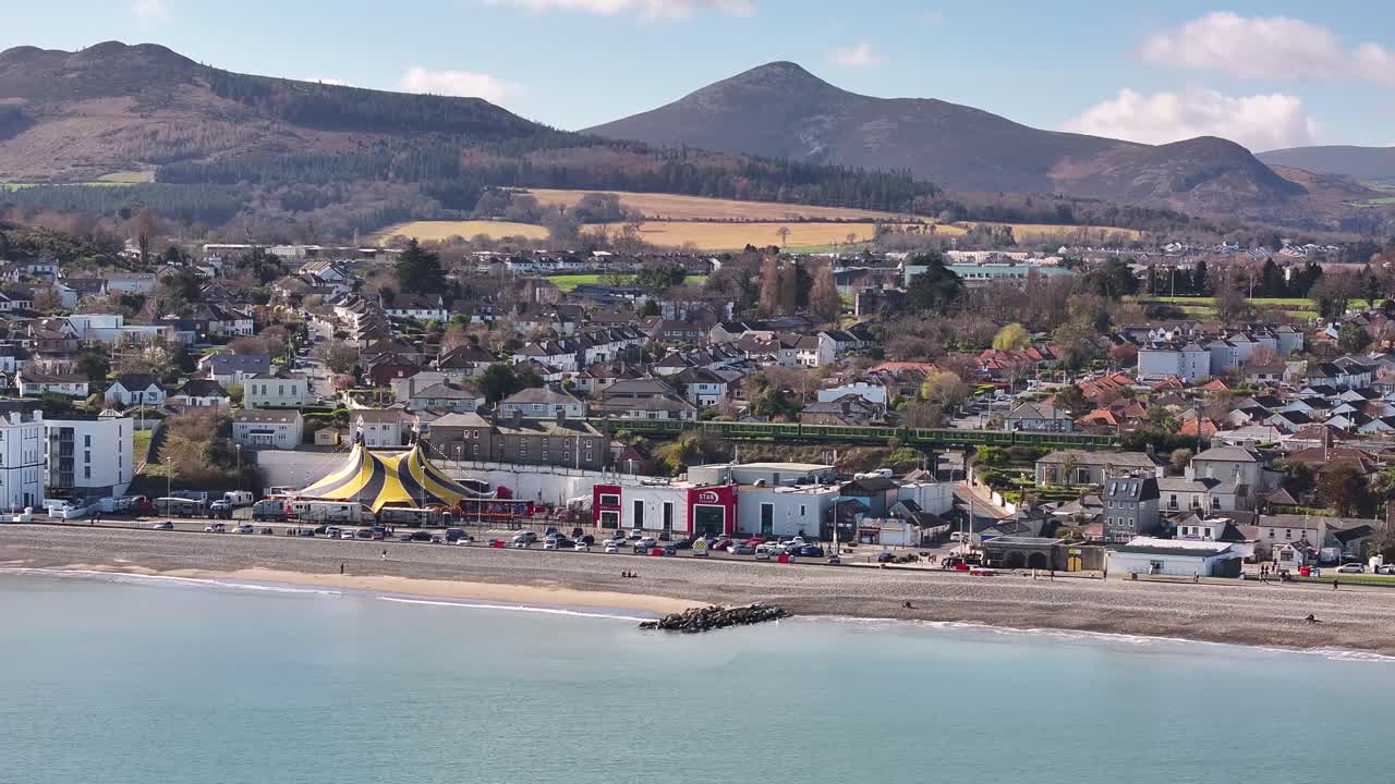 Vlak projíždí pobřežním městem Bray, letecký záběr nad pláží, v pozadí pohoří Wicklow, Irsko. Záběr dronem.