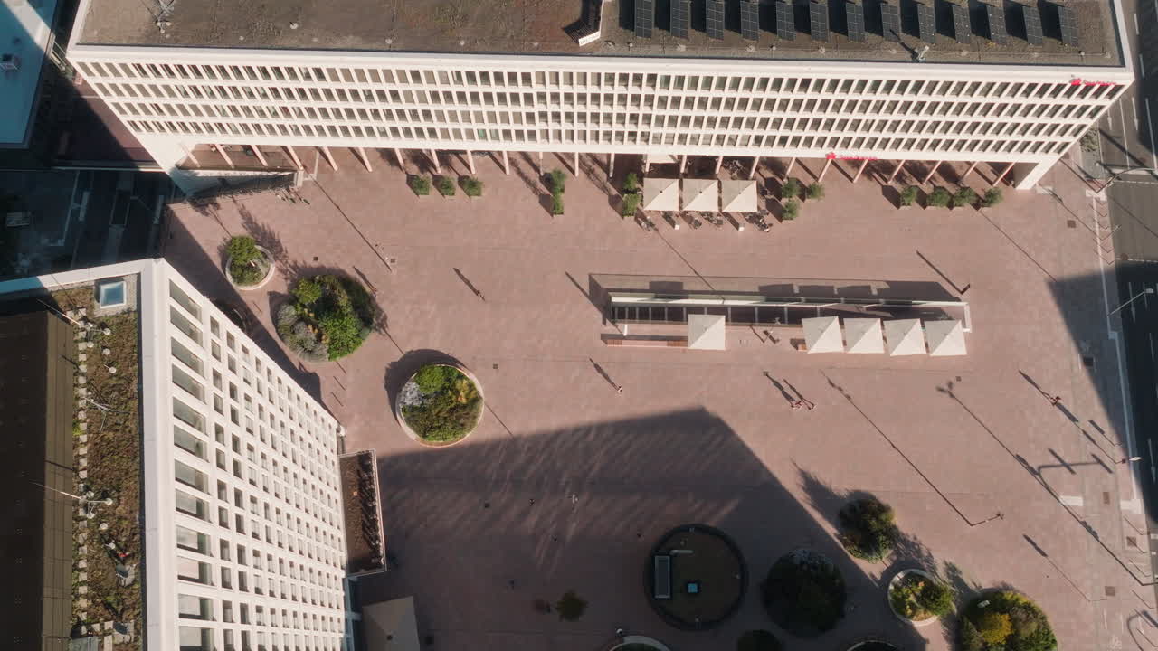 Top-down drone shot over Bahnstadt Heidelberg plaza with modern white buildings.The drone performs a slight rotation while revealing geometric buildings, paved open space and arranged trees below