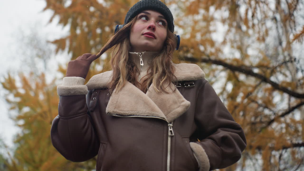 Dreamy lady wearing black knit cap and brown shearling jacket, one hand in pocket, other hand rolling hair outdoors during cool autumn day