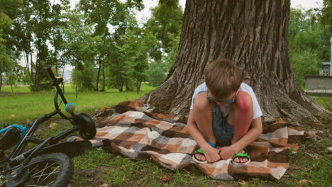 a young child sits under a large tree, deep in thought with a visible bruise on his leg, his hands are clasped together, and a blue bicycle lies on the ground nearby