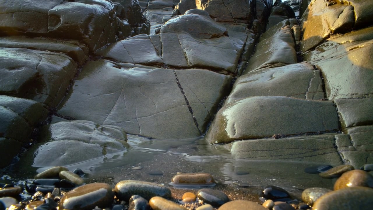 Moving swash flowing across pebbles onto bedrock cracks at shore from small wave, sunlight glinting