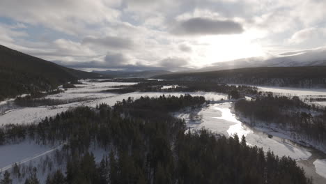 Sideways flying droneshot over a snowy valley in Norway with clouds in the sky and a low hanging sun near sunrise with trees and rivers underneath LOG