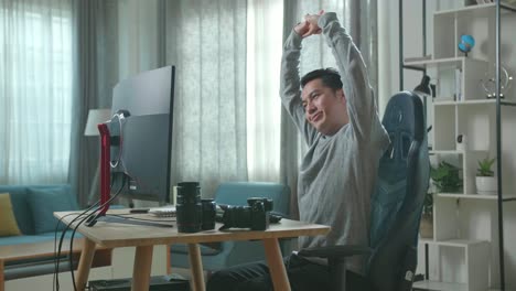 un camarógrafo asiático con una camiseta de mangas largas y pantalones negros se estira y se inclina hacia atrás mientras usa una computadora de escritorio para trabajar en casa.