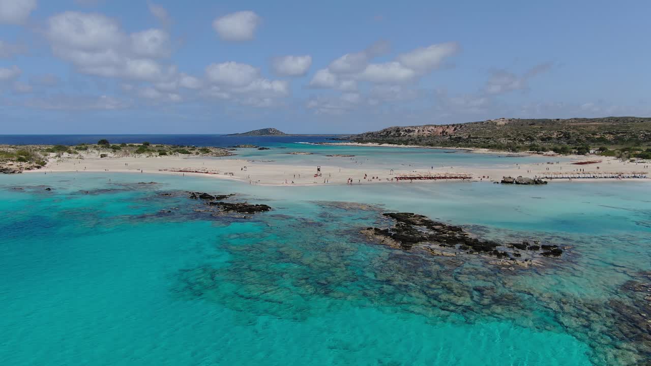 Elafonissi Beach in Crete Greece with rocks below transparent azure waters as bathers enjoy further away, Aerial hovering shot