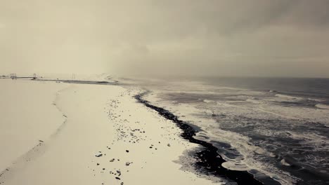 Filmische-Drohnenbewegungen-Zeigen-Einen-Schwarzen-Sandstrand-In-Island,-Der-Von-Einer-Drohne-Bei-Schnee-Gefilmt-Wurde