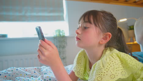 Chica-Sonriente-En-El-Dormitorio-Acostada-En-La-Cama-Usando-El-Teléfono-Móvil