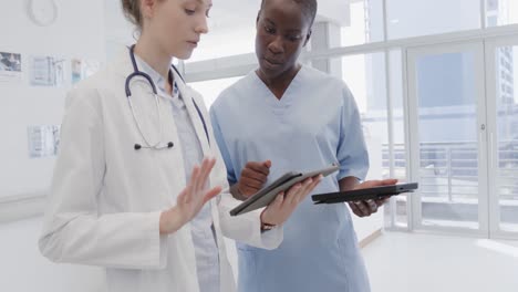 Diverse-female-doctor-and-nurse-using-tablets-and-discussing-in-hospital-corridor,-in-slow-motion