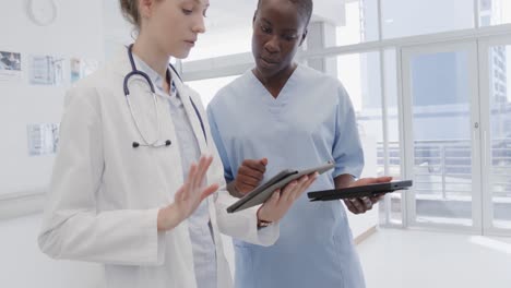 diverse female doctor and nurse using tablets and discussing in hospital corridor, in slow motion