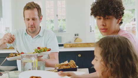 father serving as multi-racial family sitting around table in kitchen at home eating meal together