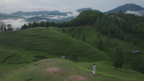 Drone-shot-of-male-tourist-walking-through-the-lush-green-hills-of-Makwanpur,-Nepal,-enjoying-raises-hands-in-celebration-amidst-vibrant-greenery-and-trees,-serene-environment-after-monsoon-rain