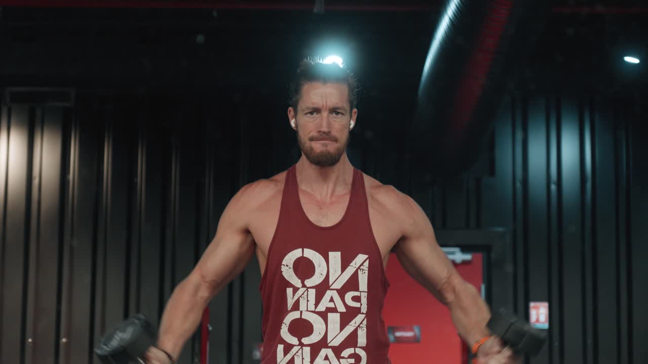 Culturista haciendo elevaciones laterales con mancuernas mientras escucha música