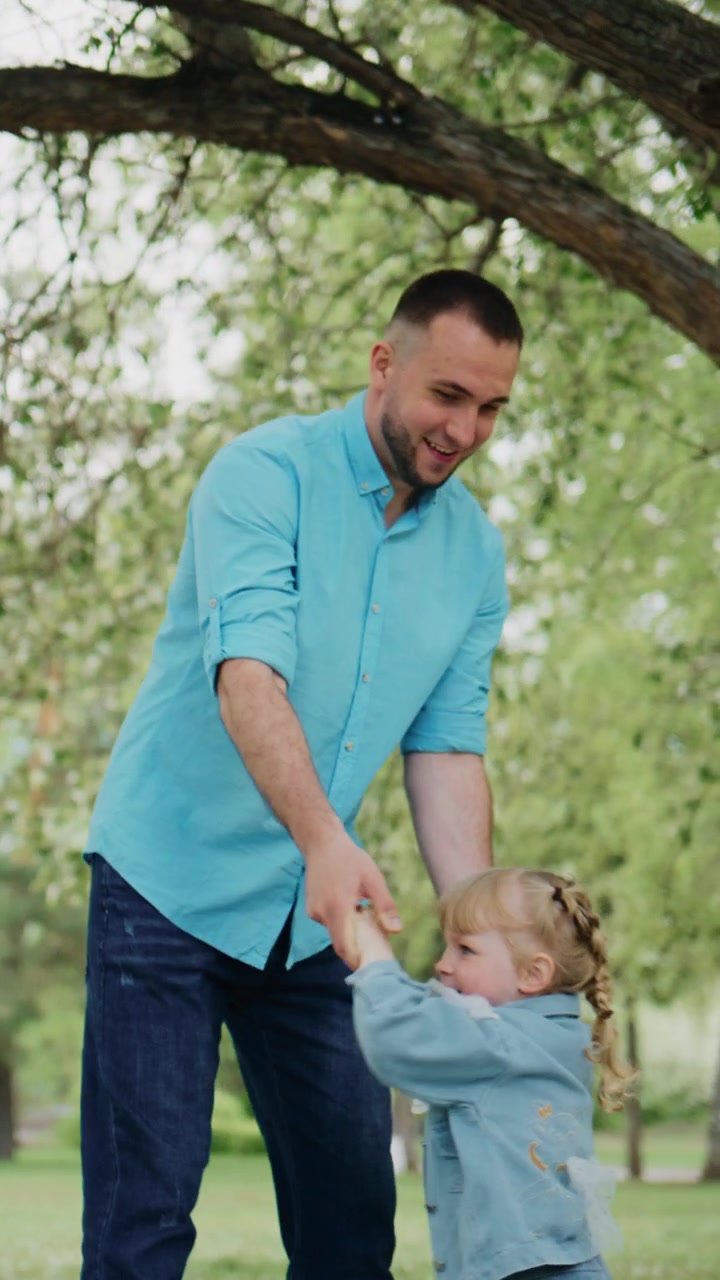 Un uomo gioca gioiosamente con la sua giovane figlia sotto un albero. La solleva delicatamente, sorridendo calorosamente mentre lei ride. La bambina gli tiene strette le mani, il suo viso pieno di pura delizia.