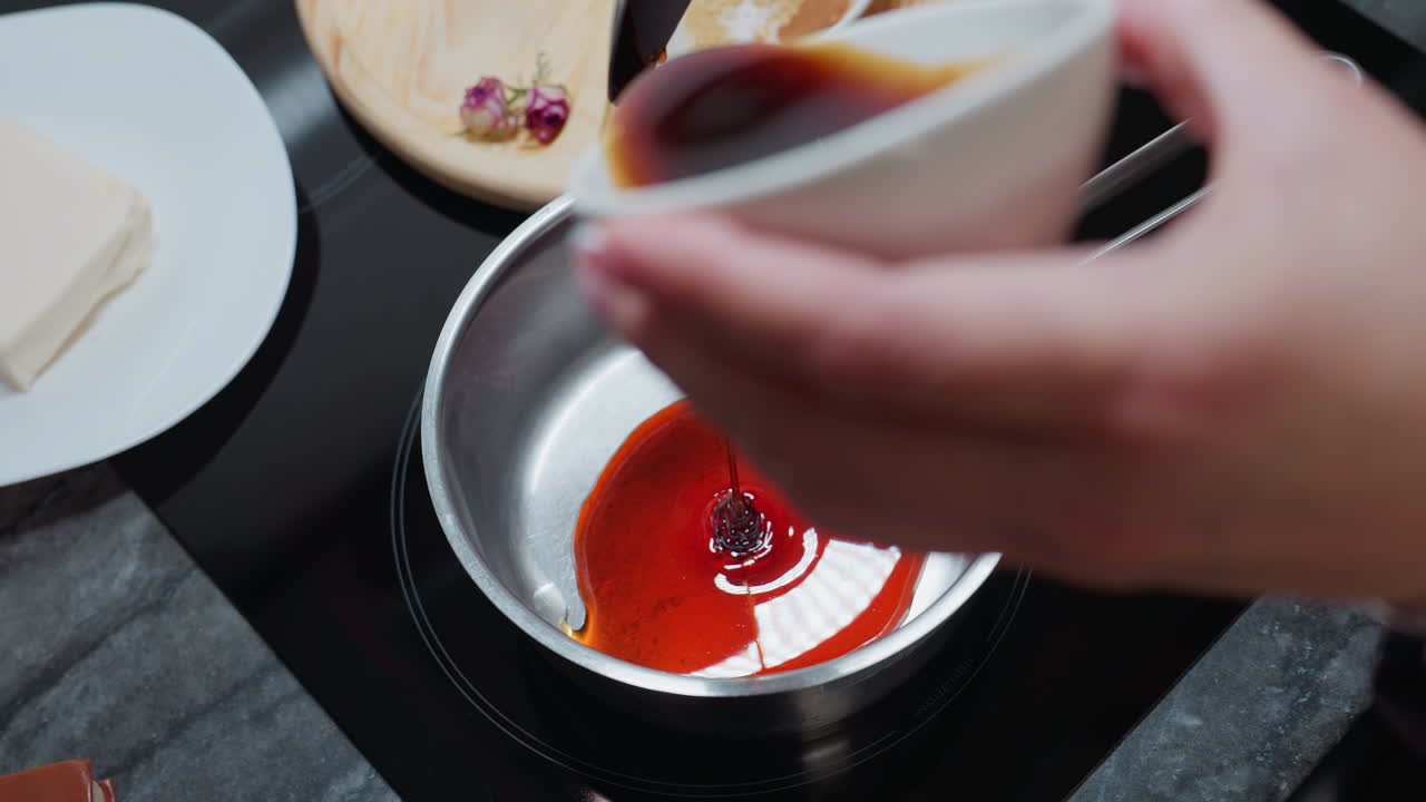 Close-up hand view of person carefully pouring honey from small white bowl into pot on electric stove, kitchen countertop with ingredients in background, warm lighting creating cozy atmosphere