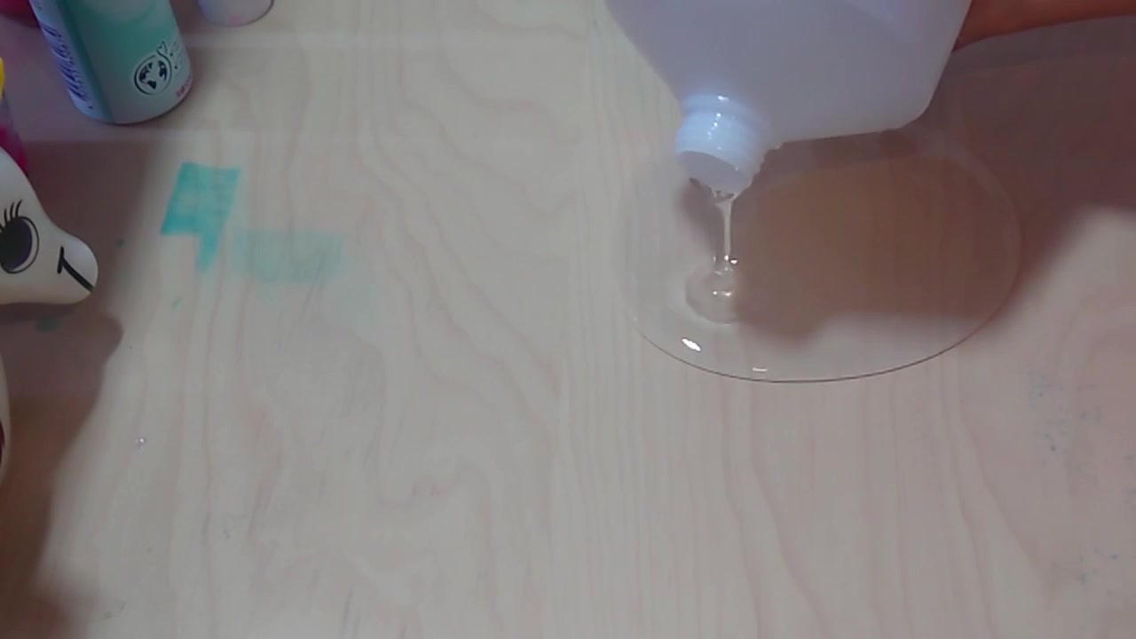 Uma tomada aérea de ângulo alto mostra cola líquida PVA transparente sendo derramada de uma garrafa plástica branca sobre uma superfície lisa de uma mesa de madeira para criar uma poça base durante um divertido experimento científico de artesanato STEM em casa.