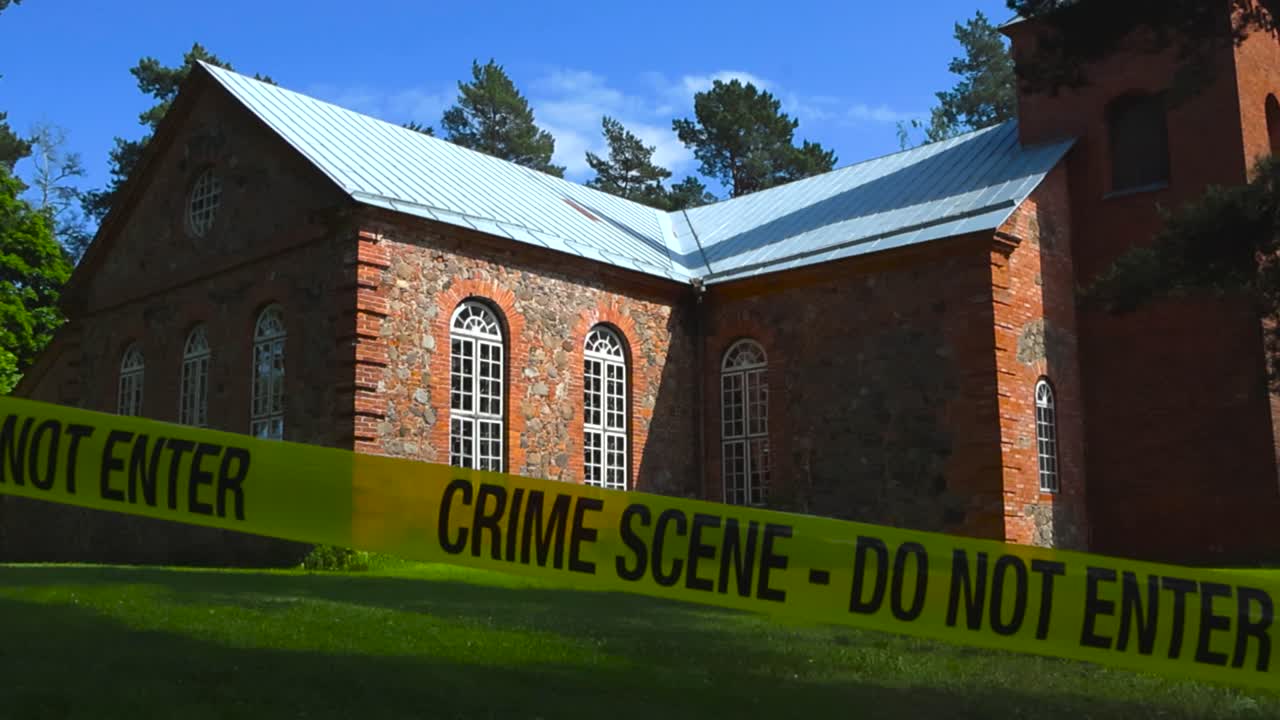 Eerie footage of a yellow vibrant colored Crime Scene Do Not Enter tape in front of an old and big red colored church in a countryside sunny forest showing a religious crime must have been commited.