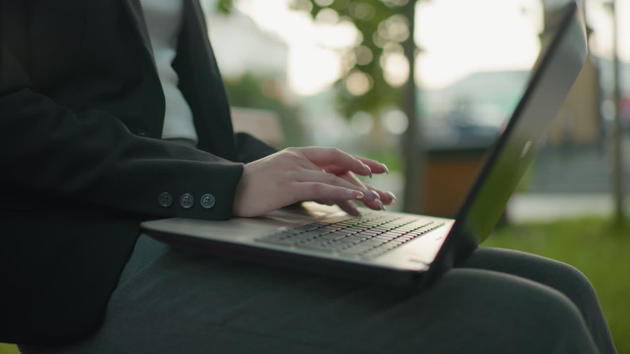 Close up side view of typist in official wear typing on laptop with polished nails in outdoor setting, background features greenery and blurred structures