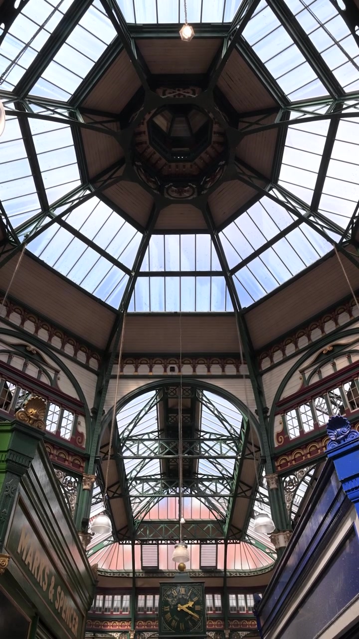 Inquadratura verticale verso l'alto del tetto in vetro ottagonale ornato e dell'orologio sospeso all'interno della galleria del Leeds Kirkgate Market.