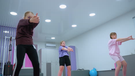 group of children in gym setting engaged in workout session, stretching arms forward with focus, background features fitness equipment, lockers, and exercise balls