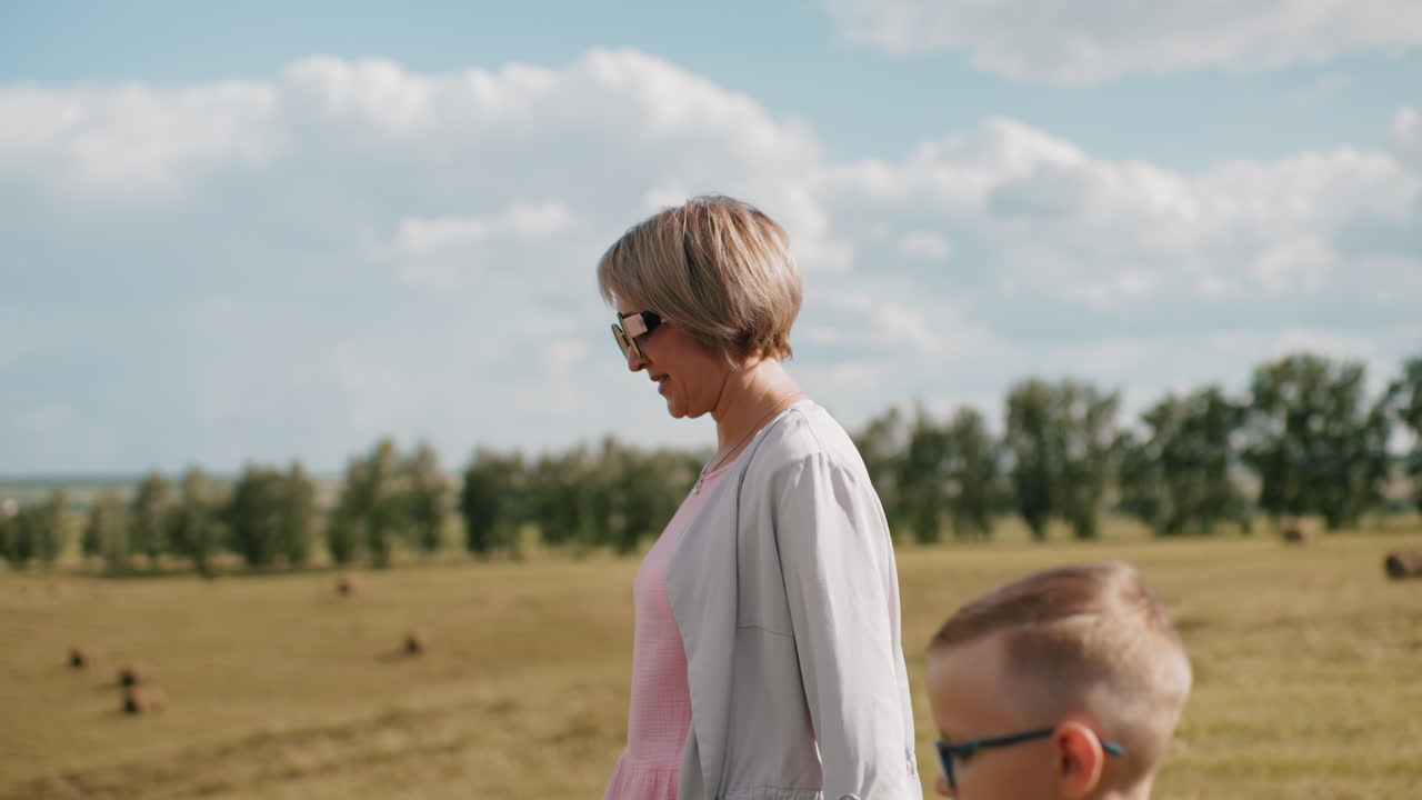 선글라스를 착용한 중년 여성이 경치 좋은 시골을 걸어다니며 줄을 들고, 안경을 쓴 손자가 동행하고, 둘 다 조용한 야외 환경을 즐기고 있습니다.