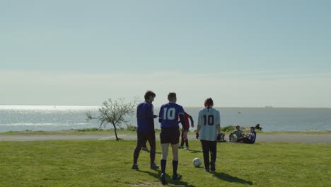two friends kick a football soccer ball near the water while others sit in the grass on a sunny day