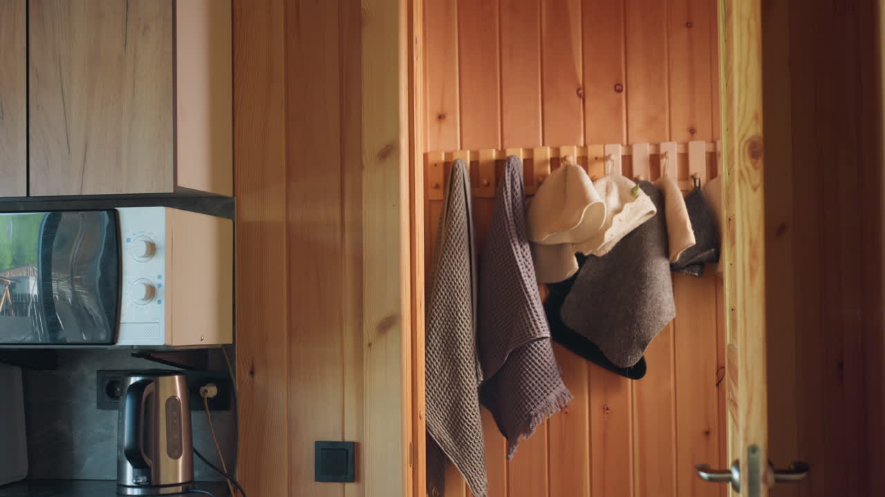 wooden sauna corner with hook rail holding waffle towels and felt hats beside microwave and kettle, warm daylight creating cosy rustic mood, textures blend pine panels with grey stone accents