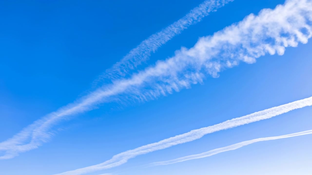 Klar blå himmel fyldt med flere hvide kondensstriber, der fejer diagonalt, herunder en tyk, fjeragtig stribe og tyndere parallelle spor, under stærkt dagslys med let dis og en jævn farvegradient.