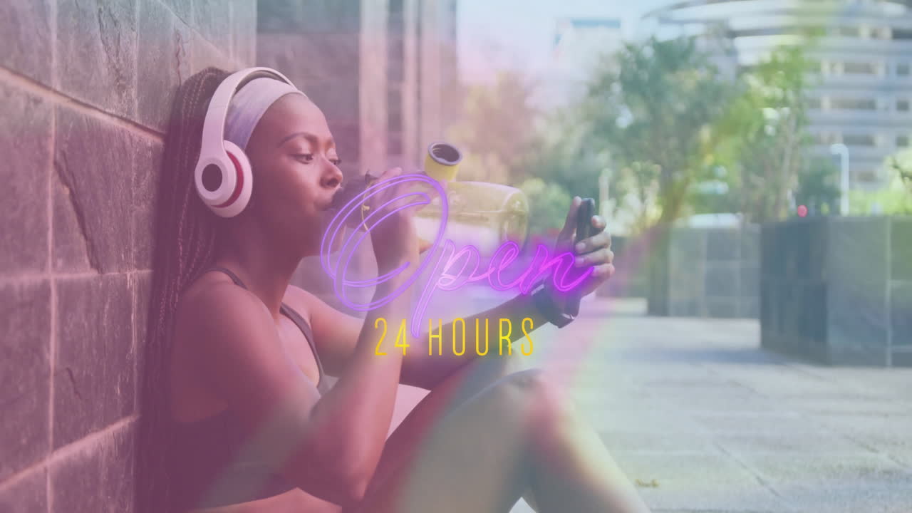 Woman leaning on brick wall, drinking water and checking fitness app, showing neon Open 24 text