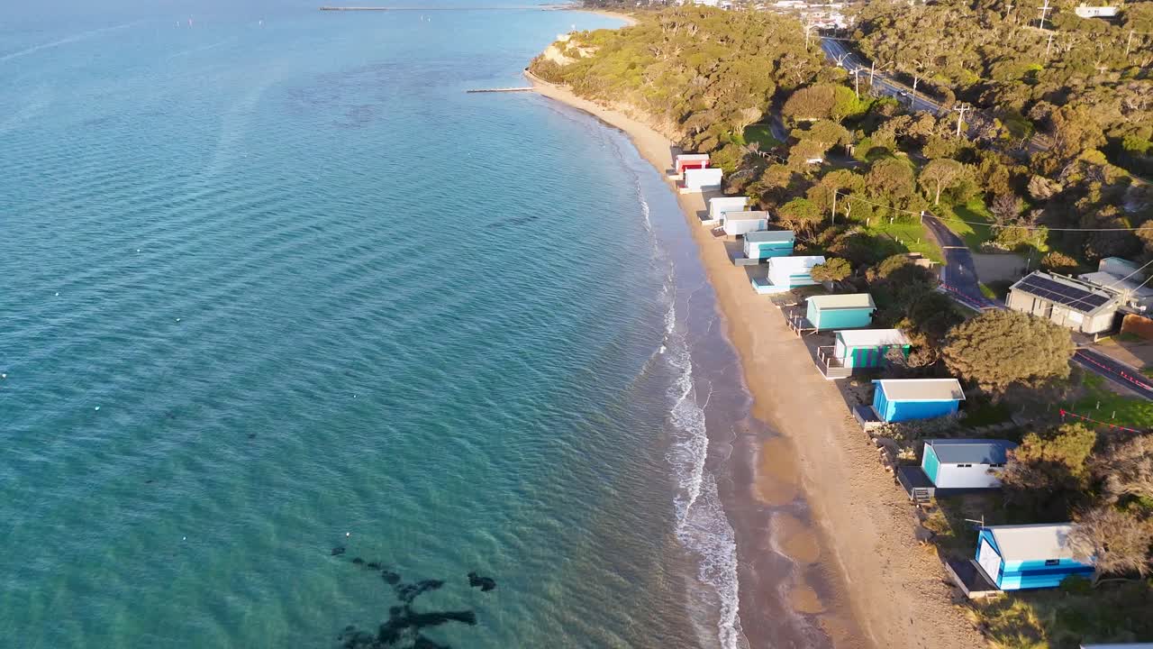 Drone glides above sandy beach, vibrant bathing boxes, and clear water in bright morning sunlight